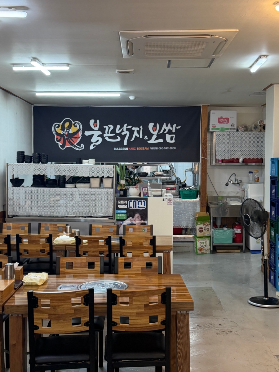 불판이 놓인 나무 테이블과 주방이 보이는 불끈낙지보쌈 화봉점 매장 내부